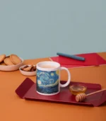 Van Gogh, Yıldızlı Gece Tasarımlı Kupa Bardak kırmızı bir tepside bal kaşığı, küçük bir bal kasesi ve çayla birlikte duruyor. Yanında kurabiye ve kuruyemiş dolu kaseler, turuncu masada defterler ve kalem yer alıyor.
