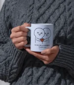 Gri, kablo örgülü kazak giyen bir kişi elinde "İçten Bir Sarılma & İsme Özel Kupa Bardak "ı tutuyor - üzerinde sarılan iki çizgi film karakteri, bir kalp ve illüstrasyonun altında Türkçe bir metin bulunan beyaz bir kupa.